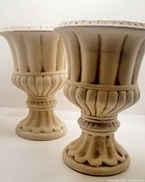 Two ceramic planters with flared tops and pedestal bases, cream color with dark distressed detailing shown at an angle