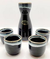 Full set of black ceramic sake bottle and four matching cups with glossy glaze and Utsuwa Japan label visible on bottle.