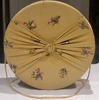 Front view of the round hat box showing beige linen-cotton fabric with floral and fruit print, pleated rosette in center, and braided handle.