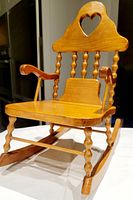 Side angle showing wooden doll rocking chair, heart cutout on backrest, curved armrests, turned spindles, rocker base, and detailed wood grain