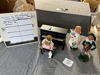 Photo showing the three figurines: Marie in pink dress sitting on piano bench, Louise seated at a black piano, and Fritz holding a nutcracker; note of the bench needing repair shown on sheet.