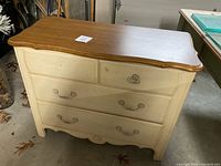 Front view of the chest of drawers showing the country white paint, three drawers with metal handles, wood top with scalloped edges, and some missing hardware.