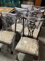 Set of six wooden dining chairs wrapped in plastic with beige upholstered seats with a damask floral pattern and carved wooden backs.