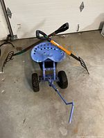 Blue gardening stool wagon with two garden pitchforks laid across the seat and front area, photographed on garage concrete floor.