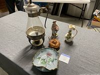 Lot of assorted vintage items on table showing coffee warmer, candy dish, figurine, jug, and lidded container