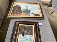 Two framed oil paintings displayed on table, larger maritime scene and smaller winter scene.