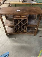 Front view of rustic wooden bookshelf showing open shelves on sides, central X-grid wine bottle storage, and drawer at top.