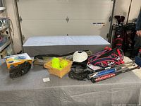 Image showing the sports gear including softball bats, helmet, glove, Adidas bag, basket of yellow softballs, badminton set and snow tube on a table in a garage environment.