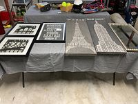 Five framed wall art pieces on table including Boston city typographic print, Paris and New York skyscraper panels, and two bridge architecture prints.
