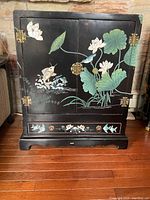 Front view of the black lacquered cabinet with hand-painted lotus flowers, birds, and fish motifs. Cabinet has ornate metal hardware on the doors and a front bottom drawer.