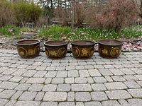 Front view of four round brown glazed planters in a row on paved surface with garden background
