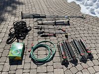 Photo displaying entire lot with garden hose, sprinklers, poles, rope, and boxed hose hanger outside on paved surface.
