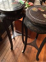Two black lacquer pedestal tables with floral bird paintings on round tops shown from a top angled side view, one with leg wear.