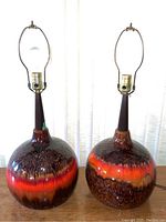 Pair of unshaded Mid Century Modern ceramic lamps with textured multicolor glaze in shades of brown, orange, and red on spherical bases with tall narrow wooden necks and brass lamp sockets.