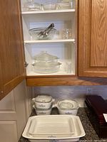Set of Corningware white ceramic baking dishes with blue floral pattern, including rectangular, square dishes, some with lids, arranged on kitchen counter and shelf.
