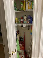 Image showing the open closet door with visible cleaning tools such as dusters, mops, brooms stored upright and some cleaning bottles on shelving.