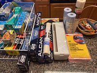 Various sizes of ALCAN aluminum foil boxes stacked with a white wrap dispenser and mesh drawer liner roll on a counter next to a pack of reusable cleaning cloths, bubble wrap rolls, and storage shelf.