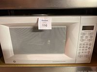 Front view of GE microwave oven showing door with window and mesh screen, and MaxSold lot number tag.
