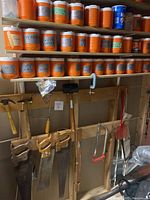 Wide shot showing rows of labeled orange containers and a few blue containers with nuts and bolts on shelves above several hand saws mounted on a wooden rack.