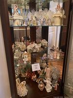 Photo showing multiple angel figurines displayed on glass shelves inside a wooden cabinet, including porcelain angels, ceramic angels, and some floral-themed angel pieces.