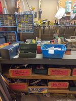 Wide view of a workbench with multiple labeled cardboard boxes containing shelf brackets, hooks, hangers, wires, and cables. Several metal toolboxes and plastic organizers with small hardware under and on top of the bench.