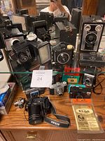 Wide view of the collection of vintage cameras arranged on and in front of a mirror, showing multiple box cameras, old film models, and a Nikon digital camera with strap.