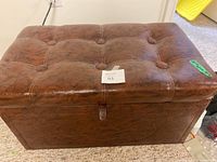 Full view of the brown leather tufted trunk showing rectangular shape, button tufting on top, and pull tab for opening.
