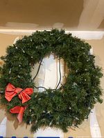 Green artificial evergreen wreath hanging against a wall with white string lights wrapped around it and a red decorative bow attached.
