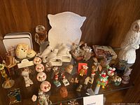 Wide view showing various owl figurines on glass shelf including ceramic, glass, metal, and painted owls, large white ceramic owl plate/tray, and grouped owl pins and coaster set.