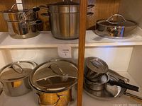 Full view of multiple stainless steel pots and pans with lids including saucepans, frying pans and pasta strainer
