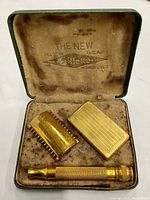 Top-down image of the gold tone razor set including the handle, razor head, and blade case, all laid in the original velvet lined box with printed logo inside lid.