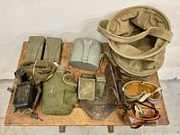 Photo of whole lot on wooden table showing assorted military gear including canteen with cover, canvas bag, goggles, leather holster, belts, metal mess kit, and pouches.