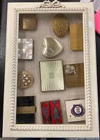 All eleven vintage compacts arranged inside a cream carved wooden display case with glass top, showing diverse designs including mother-of-pearl, enamel, and gold-tone metal finishes