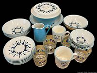 Display of multiple Marcrest Swiss Chalet pattern plates and bowls alongside Blue Heaven solid blue pitcher, covered bowl with replacement lid, six decorated drinking glasses and two mugs with floral patterns.