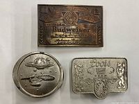 Three vintage metal belt buckles shown from front: Budweiser rectangular, Coors rectangular, Star Trek oval spaceship design.