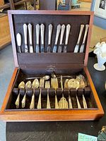 Opened wooden silverware box displaying the set of utensils with the top row showing knives and butter spreader and bottom compartment showing various spoons, forks, and tongs.