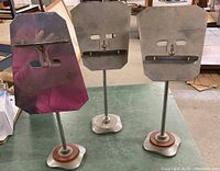 Photo showing all three metal shoestore stands upright on a green surface, displaying their front clamp plates and weighted bases.