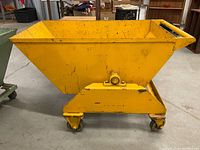 Side view of yellow industrial metal wheelbarrow hopper on caster wheels with handle and pivot mechanism.