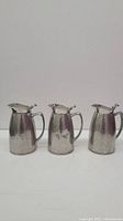 Three stainless steel milk pitchers with hinged lids and handles, shown together from the front