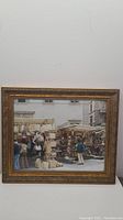 Front view of framed artwork depicting an outdoor market scene with people and stalls, set in a decorative gold frame.