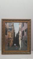 Framed artwork showing a European narrow alleyway canal with man rowing a boat, wooden carved ornate frame