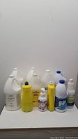 Full view of all items in lot showing four gallon jugs, yellow bottles, a bleach bottle, foaming soap dispenser and spray bottle.