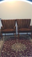 Photo showing the pair of vintage Italian armchairs side by side on a patterned rug, highlighting their scalloped backs and wooden legs.