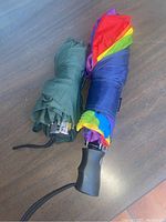 Two compact folded umbrellas, one olive green and one rainbow-colored, showing handles and wrist straps.