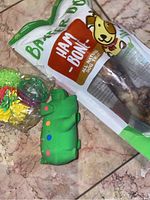 Photo of Barkworthies Ham Bone dog treat in sealed packaging along with a green pig rubber squeaky toy and a multicolor rope ball toy on floor