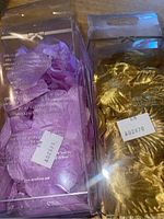 Two packs of artificial flower petals in plastic containers, one gold and one lavender, with brand name and barcodes visible.