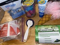 Overview of lot showing Neutrogena makeup remover wipes, gauze sponges, biodegradable trash bags, white handle brush with blue scrubbing pad, cone-shaped scrub brush with pink mesh pouf, bath bombs, and Crayola finger paint soap.