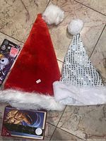 Two Christmas hats on tile floor: one red with white fur trim and pom-pom, one silver with white fur trim and black sequins