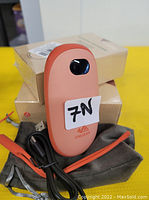 Two orange UNIGEAR rechargeable handwarmers with power buttons, USB charging cables, wrist straps, and gray sleeves on a yellow surface with product boxes in the background.
