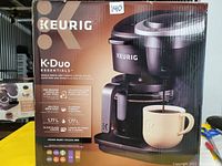 Front view of the Keurig K-Duo Essentials box showing the product image, brand, and feature highlights including 12-cup carafe, single serve, 1.77L reservoir, black color.